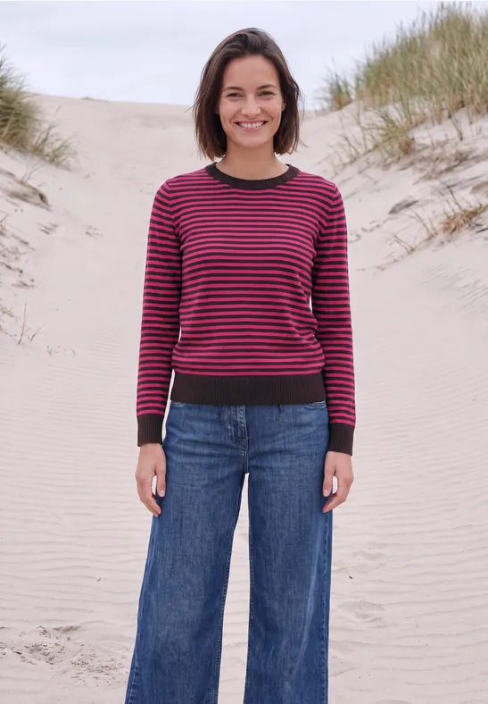 Een vrouw in spijkerbroek en gestreepte trui staat op een duin bij de zee.