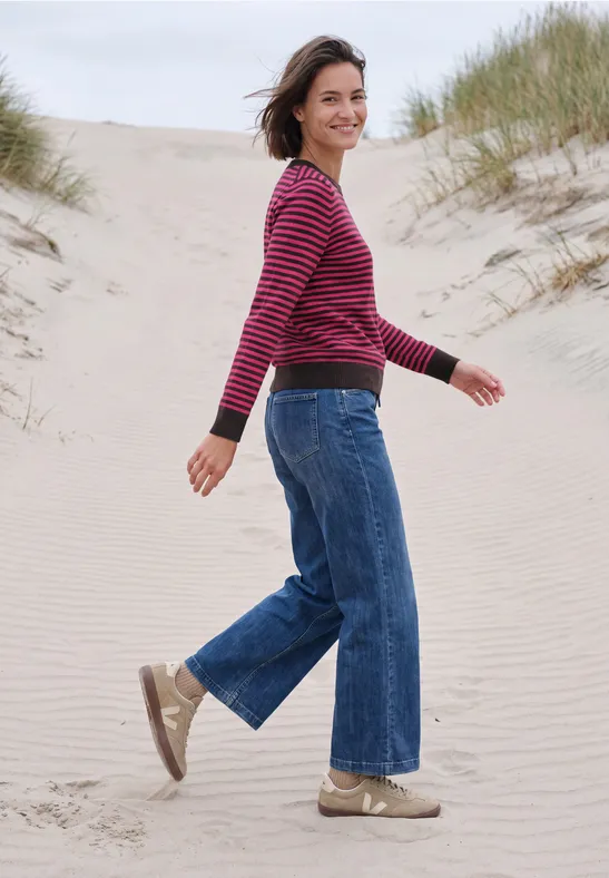 Een vrouw in een rood gestreepte trui en wijde spijkerbroek loopt lachend over zandduinen op het strand.