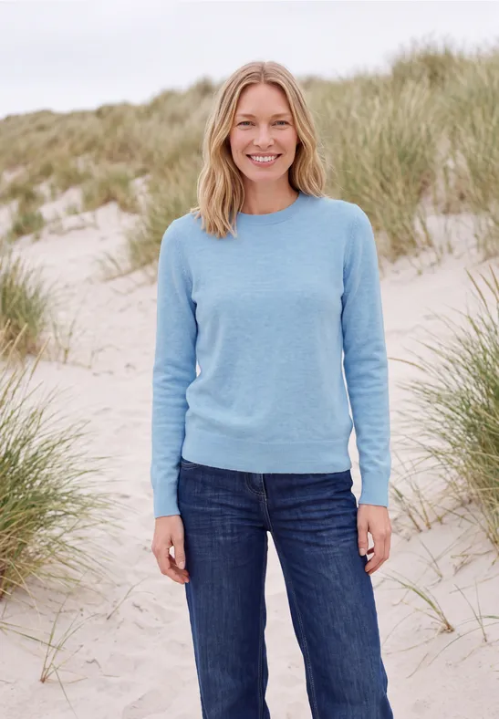 Eine Frau steht lächelnd in blauem Pullover und Jeans am Strand zwischen Dünen.