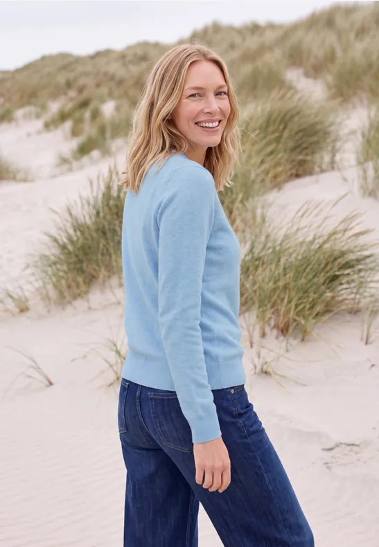 Lächelnde Frau in blauem Pullover und Jeans steht am Strand vor Dünen mit Strandhafer.