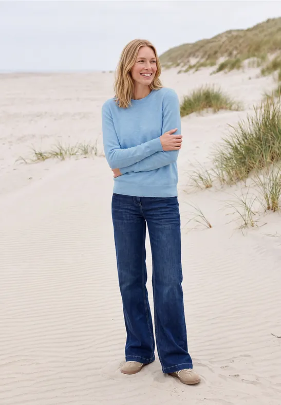 Eine Frau in blauem Pullover und Jeans steht lächelnd am Strand vor Dünen.