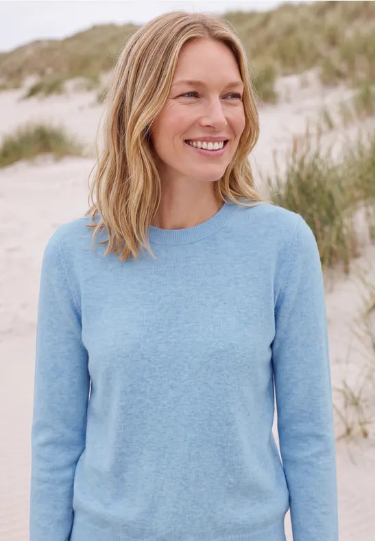 Lächelnde Frau in hellblauem Pullover steht an einem Dünenstrand.