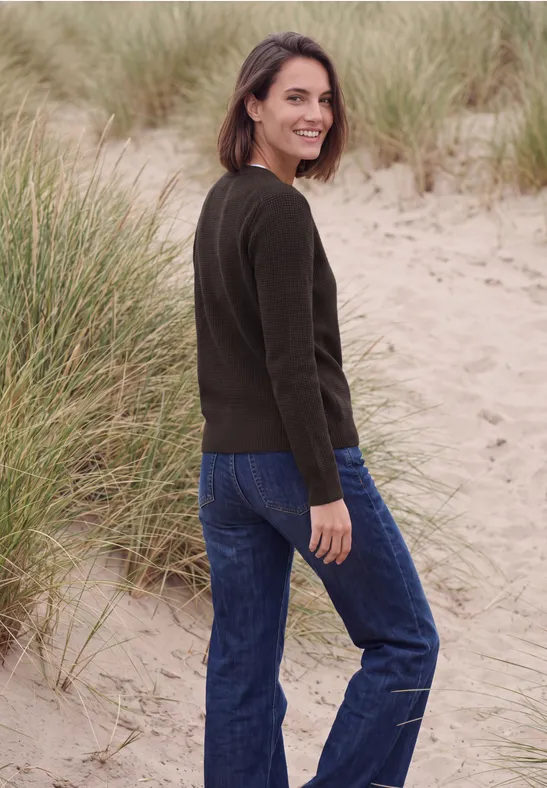 Vrouw in groene trui en spijkerbroek staat in duinen, kijkt met een glimlach over haar schouder.