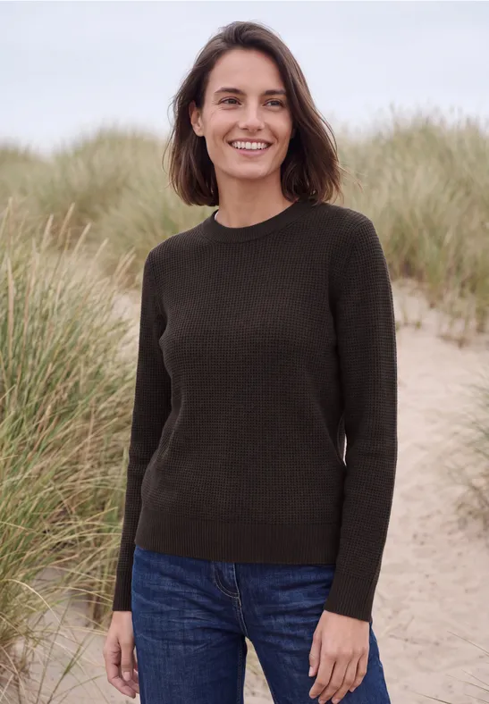 Een glimlachende vrouw staat in duinen voor hoog gras en draagt een spijkerbroek en een donker truitje.