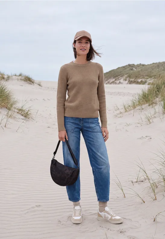 Vrouw in spijkerbroek, trui en pet met tas, lopend op zandduinen bij het strand.