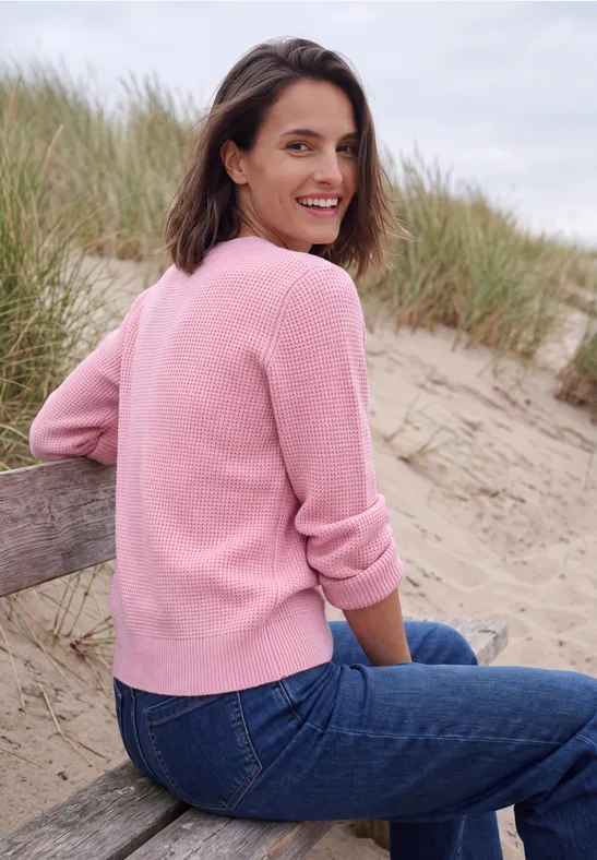Een vrouw in een roze trui en spijkerbroek zit glimlachend op een bankje in de duinen bij het strand.