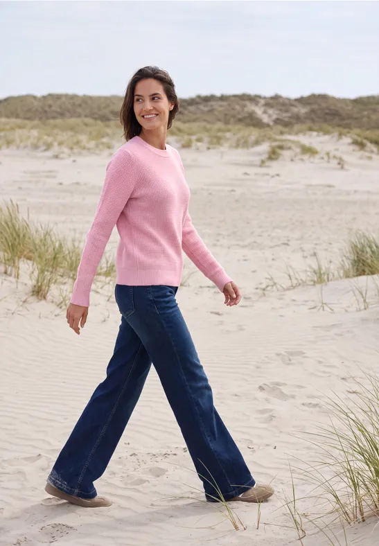 Een vrouw loopt over het strand in spijkerbroek en roze trui.