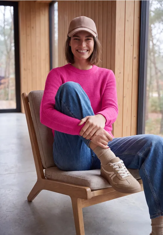 Junge Frau in rosa Pullover, Jeans und Cap sitzt lächelnd auf einem Holzstuhl in hellem Raum.