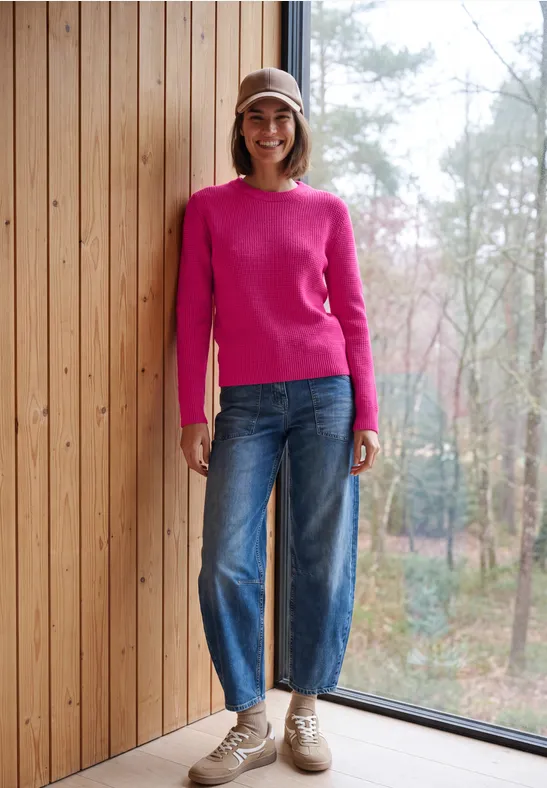 Eine lächelnde Frau in pinkem Pullover, weiten Jeans und beigem Hut lehnt an einer Holzwand vor einem Fenster.
