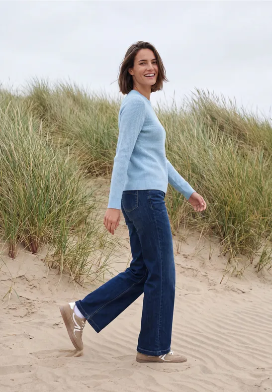 Vrouw in lichtblauwe trui en jeans loopt glimlachend over een zandstrand met helmgras op de duinen op de achtergrond.