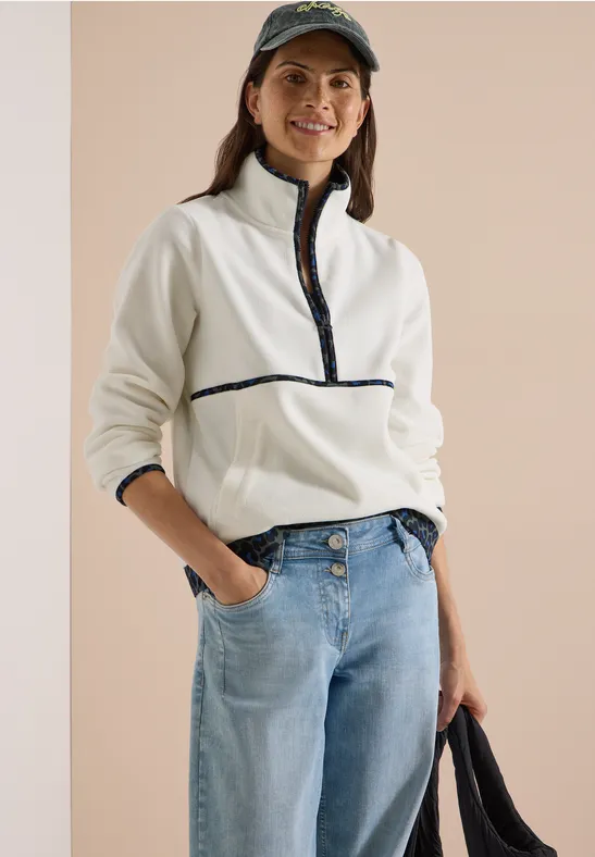 Lachende vrouw draagt een witte fleece met rits, jeans, pet en houdt een tas vast.