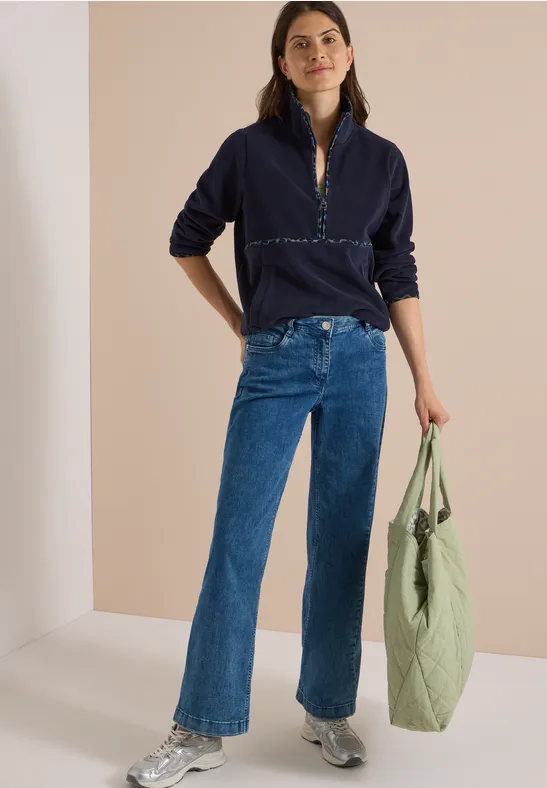 Vrouw in donkerblau trui en jeans, draagt een lichtgroene tas van stof en glimlacht ontspannen.
