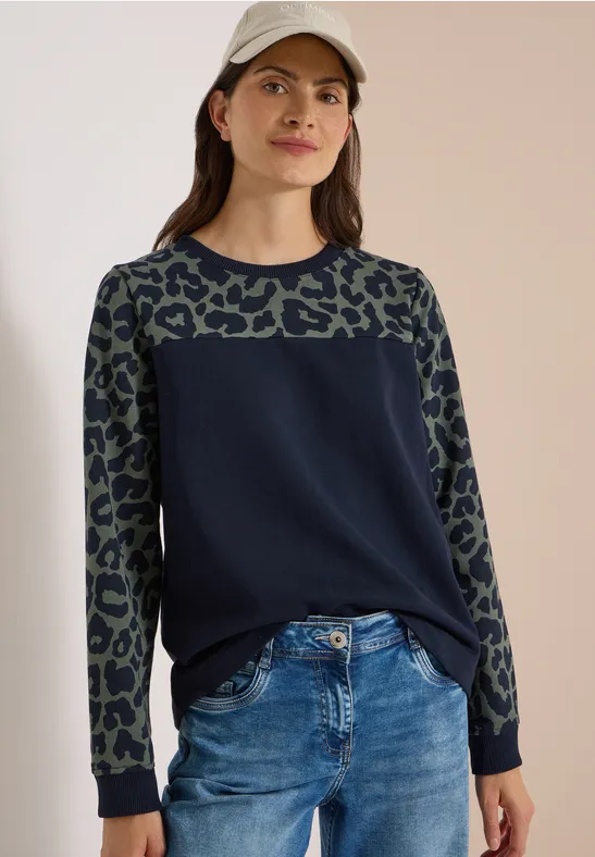 Frau in dunkelblauem Pullover mit Leopardenmuster an Ärmeln und Jeans, mit Basecap vor neutralem Hintergrund.