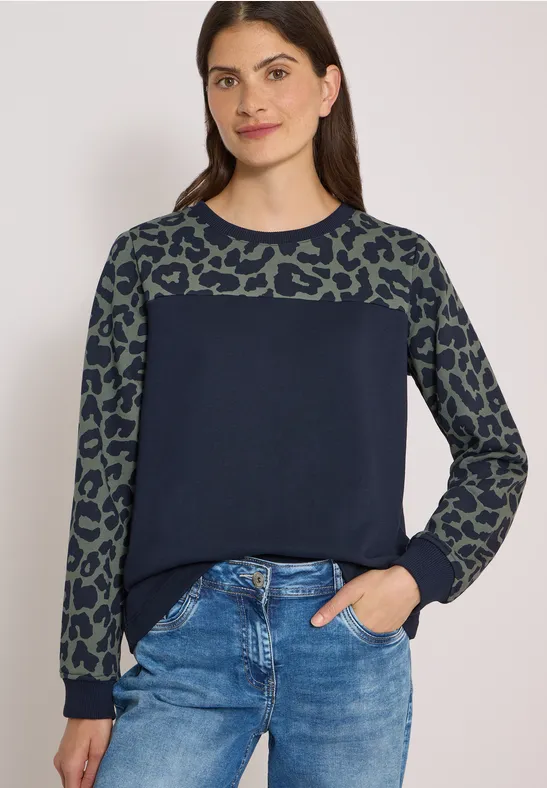 Junge Frau trägt Jeans und Pullover mit marineblauem Oberteil und grünen Leomustärmeln, Hand in Hosentasche.