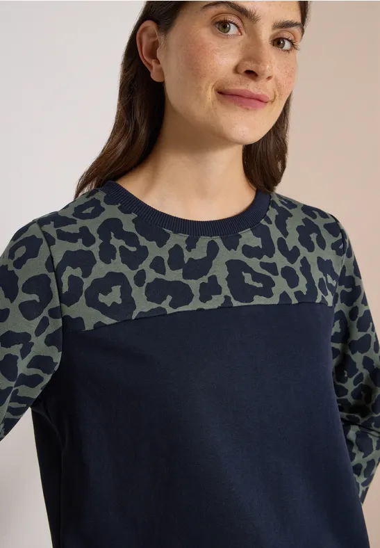 Frau im dunkelblauen Pullover mit grün-schwarzem Leopardenmuster oben und an den Ärmeln.