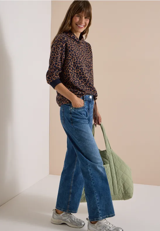 Frau mit Leo-Bluse, weitem Jeans, Sneakern und großer grüner Tasche, lächelnd seitlich in heller Umgebung.