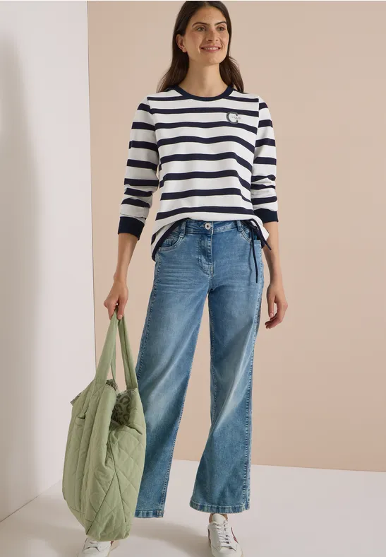 Femme en haut à rayures blanc-bleu, jean large, baskets blanches, tenant un cabas matelassé vert.