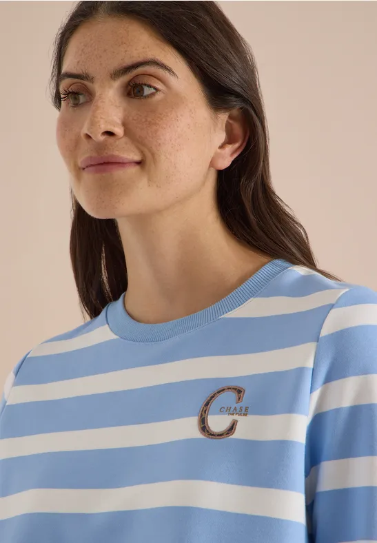 Frau trägt hellblau-weiß gestreiftes T-Shirt mit kleinem goldfarbenen 'C'-Logo auf der Brust.