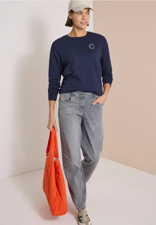 Frau in dunkelblauem Sweatshirt, grauen Jeans und Cap, hält eine orange Stepp-Tasche und trägt Sneakers.