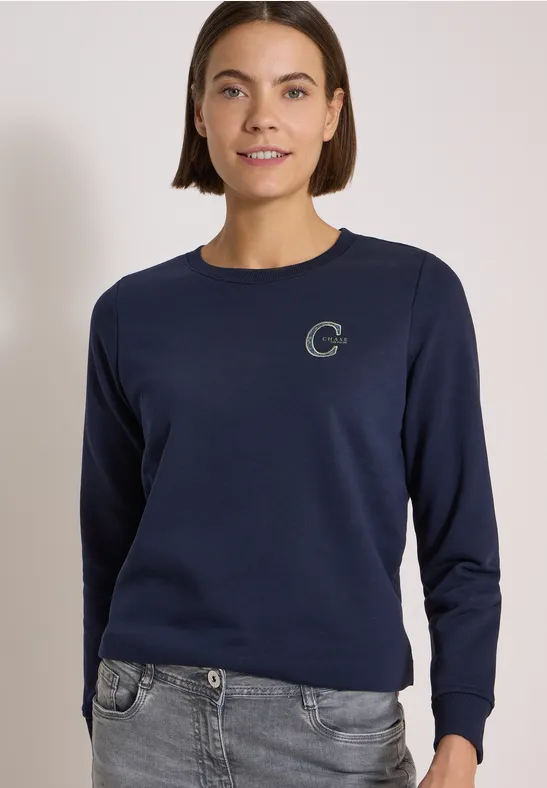Frau trägt dunkelblaues Sweatshirt mit rundem Ausschnitt und kleinem 'C'-Logo, kombiniert mit grauer Jeans.