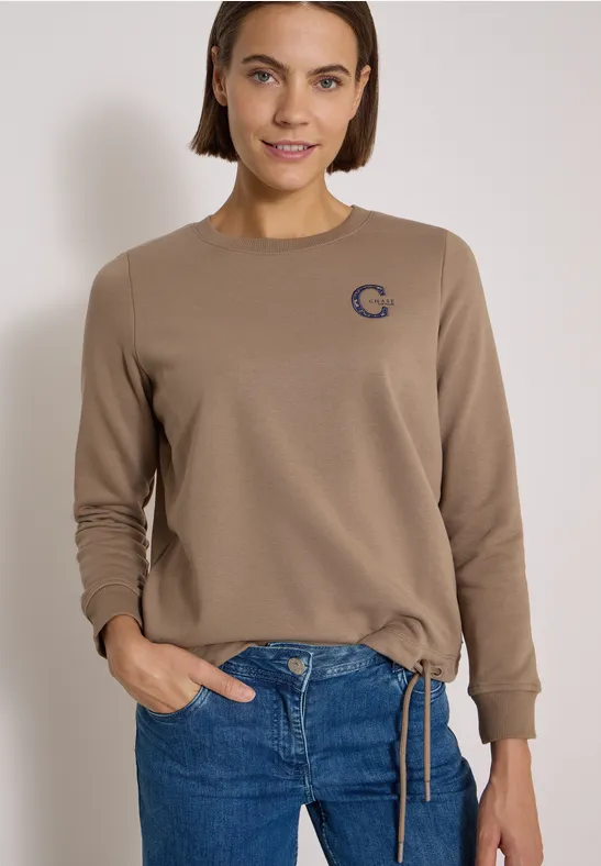 Frau trägt beigen Pullover mit kleinem Logo und blaue Jeans.