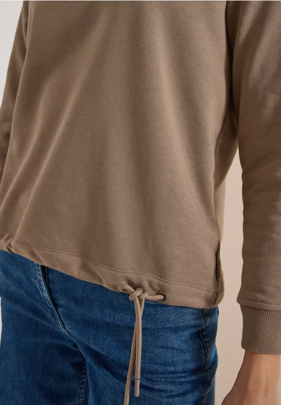 Braunes Langarmshirt mit Kordelzug am Saum, kombiniert mit blauen Jeans.