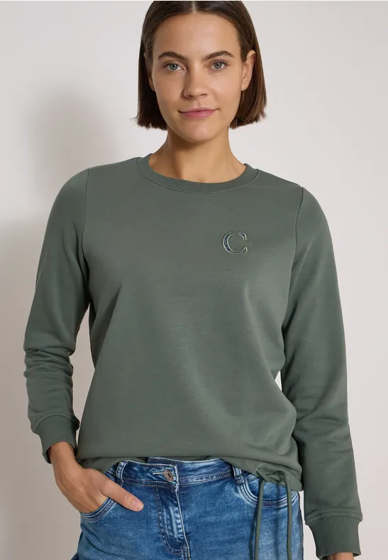 Frau trägt grünes Sweatshirt mit kleinem Logo und blaue Jeans vor hellem Hintergrund.