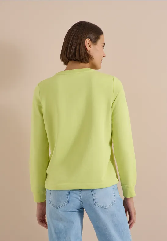 Frau von hinten in hellgrünem Langarmshirt und Jeans vor beigem Hintergrund.
