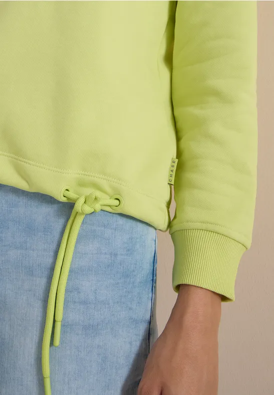 Leuchtend grüner Hoodie mit Kordelzug am Saum und hellblauer Jeans.