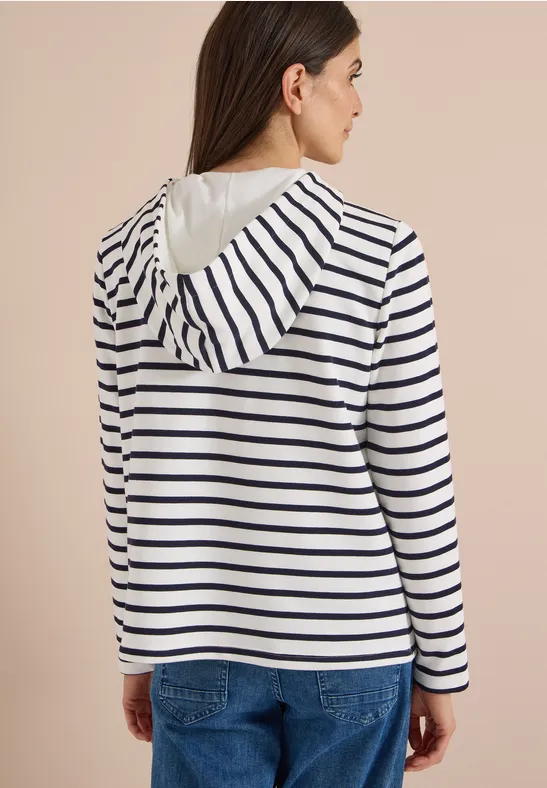 Eine Frau von hinten, trägt einen weißen Kapuzenpullover mit dunkelblauen Streifen und Jeans.