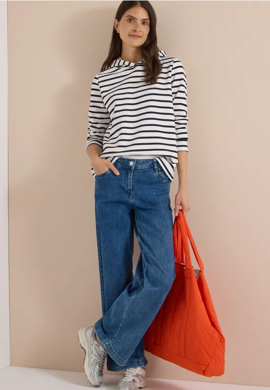 Frau in gestreiftem Langarmshirt, weitem Jeans, weißen Sneakers und orangefarbener Tasche vor neutralem Hintergrund.