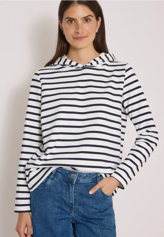Junge Frau in weiß-blau gestreiftem Hoodie, Jeans, lächelt, halb in die Tasche gesteckte Hand.