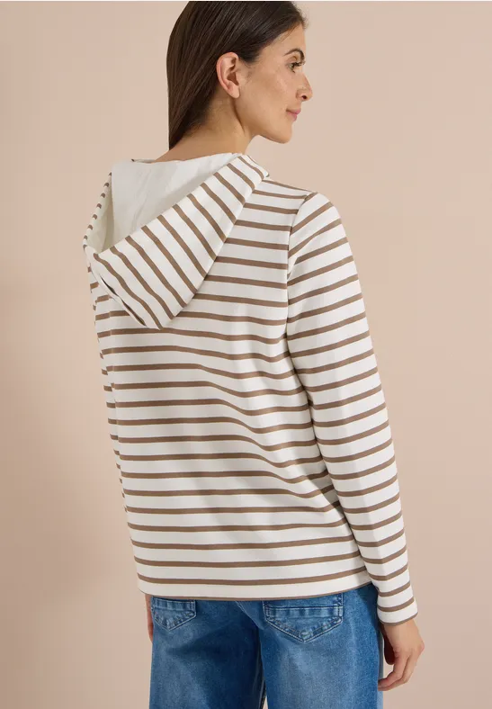 Frau trägt beige-weiß gestreiften Hoodie und blaue Jeans, seitliche Ansicht vor neutralem Hintergrund.