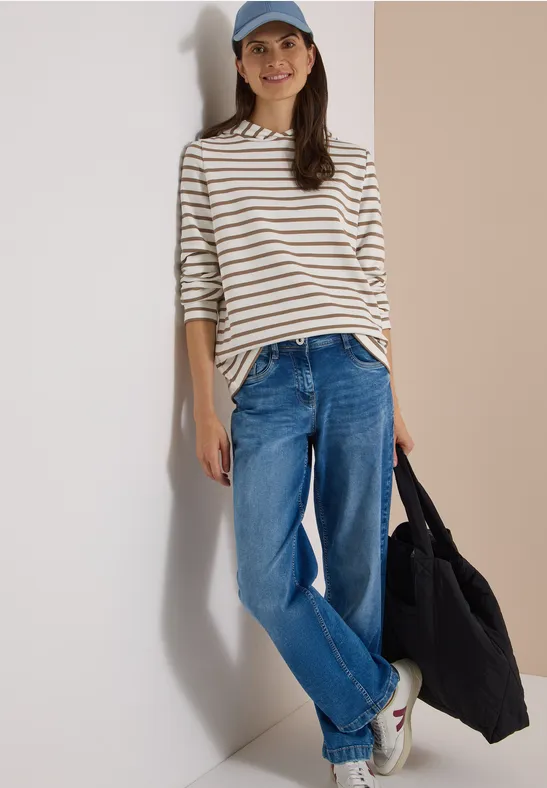 Frau lehnt an Wand, trägt gestreiftes Langarmshirt, Jeans, Baseballcap, weiße Sneaker und hält schwarzen Shopper.