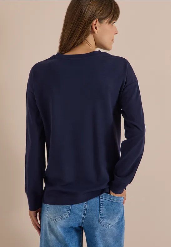 Femme de dos portant un pull à manches longues bleu marine et un jean bleu, les mains dans les poches.