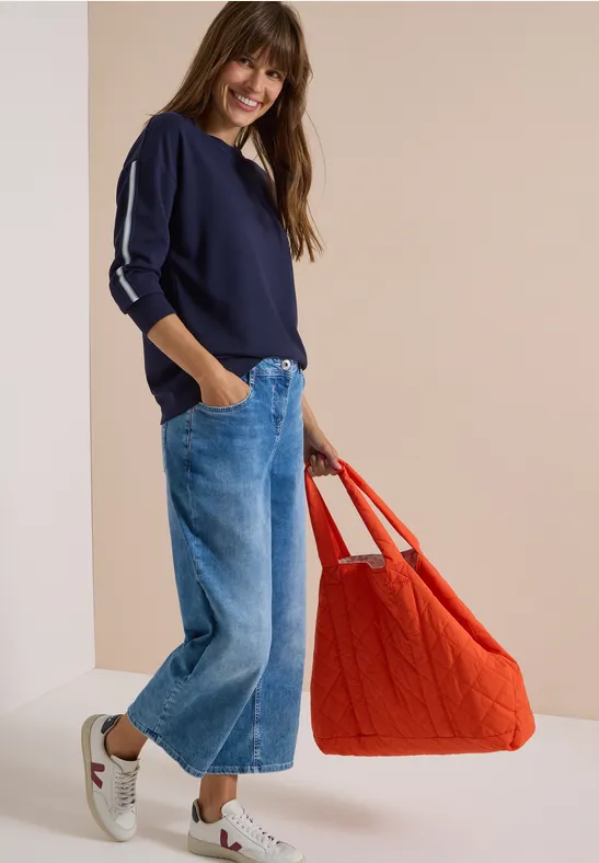 Une femme porte un pull bleu marine, un jean large, des baskets blanches et un grand sac rouge.