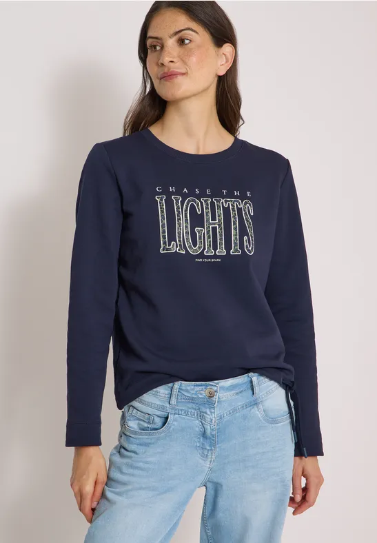 Kobieta w granatowej bluzie z napisem 'LIGHTS' i jasnych dżinsach, na białym tle.