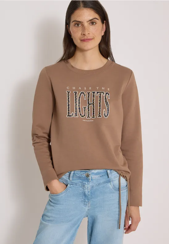 Eine Frau in braunem Sweatshirt mit Schriftzug ‘AMAZING LIGHTS’ und blauen Jeans vor hellem Hintergrund.