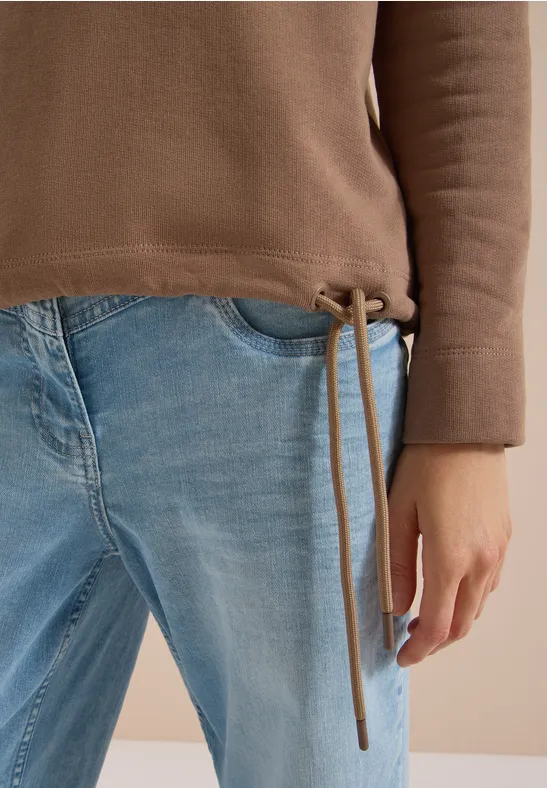 Eine Person trägt eine braune Pulloverjacke mit Tunnelzug und hellblaue Jeans; die linke Hand ruht locker an der Seite.