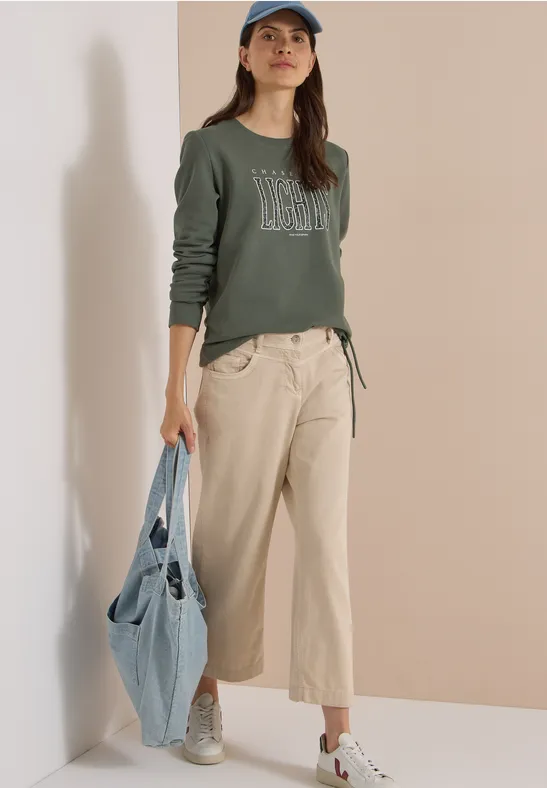 Frau in grünem Sweatshirt, beigen Hosen, weißen Sneakern und Baseballcap, hält eine Jeans-Tasche.