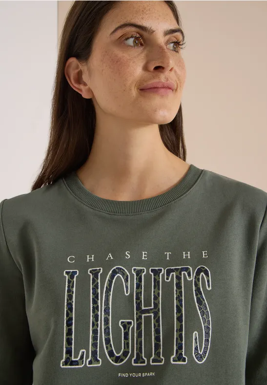 Frau mit langen dunklen Haaren trägt grünes Shirt mit Aufschrift 'CHASE THE LIGHTS'. Sie blickt seitlich.