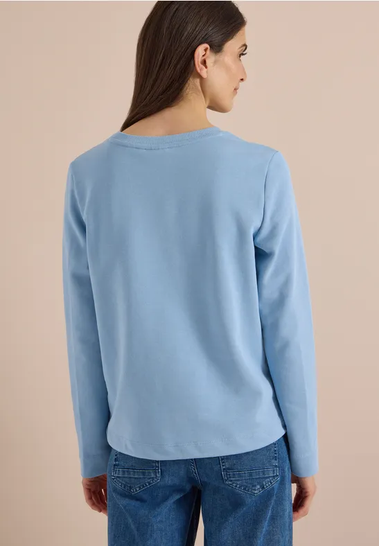 Frau von hinten in hellblauem Langarmshirt und Jeans vor neutralem Hintergrund.