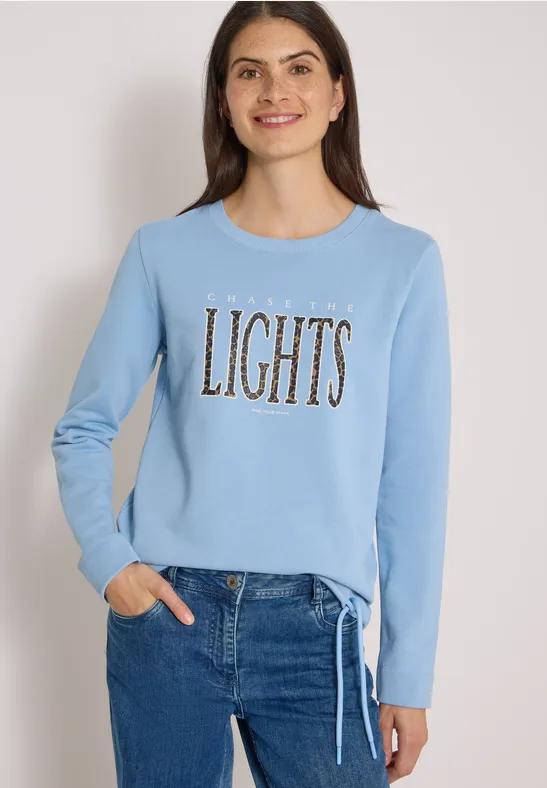 Frau im hellblauen Sweatshirt mit goldfarbenem Schriftzug "LIGHTS" und Jeans, lächelnd.