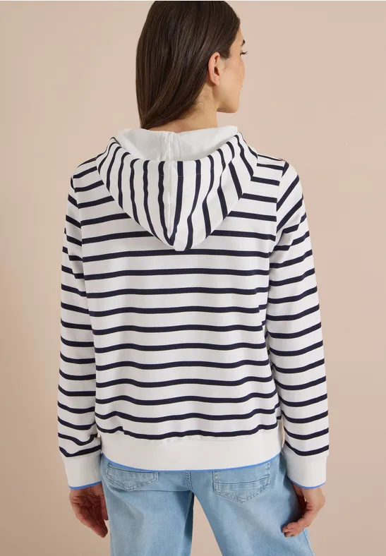Frau von hinten in weißer Kapuzenjacke mit dunkelblauen Streifen und blauen Jeans.