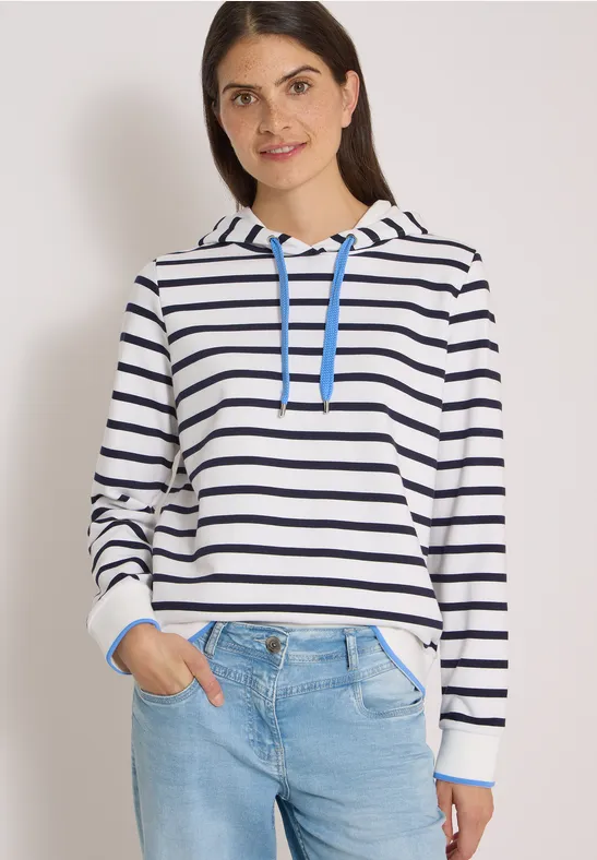 Junge Frau trägt einen weiß-blau gestreiften Kapuzenpullover und Jeans vor hellem Hintergrund.