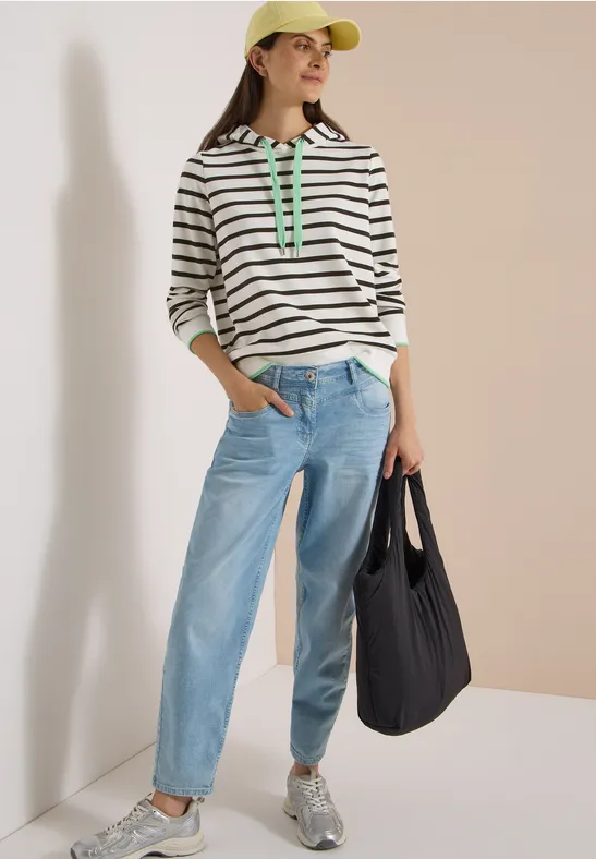 Frau in gestreiftem Hoodie, Jeans, Cap und Sneakern mit schwarzer Tasche vor hellem Hintergrund.