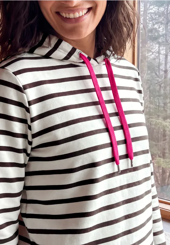 Nahaufnahme einer Frau mit gestreiftem Kapuzenpullover und pinken Kordeln, die lächelnd posiert.