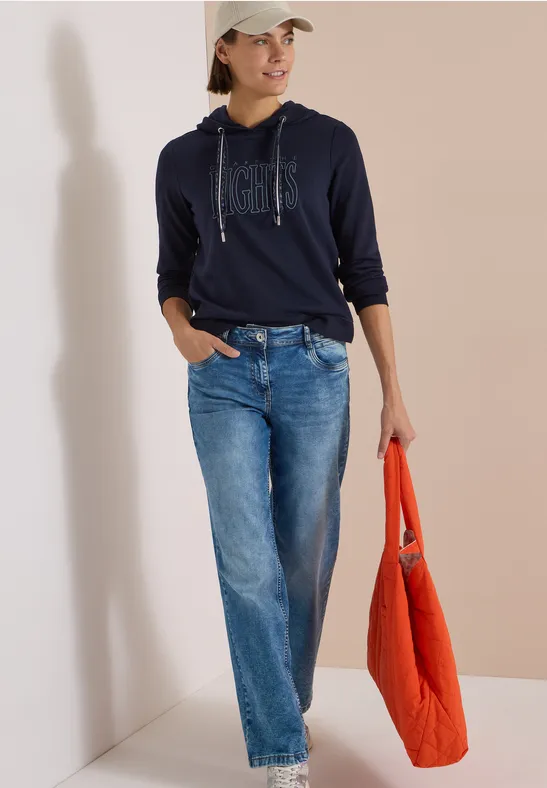Frau in Navy-Hoodie, weiten Jeans und Basecap, trägt eine orangefarbene Tasche.
