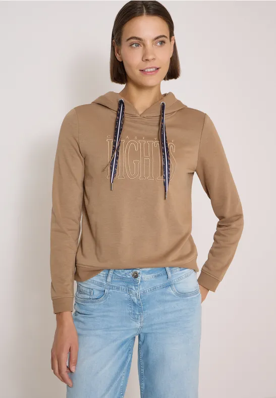Junge Frau in beigem Hoodie mit 'icon'-Aufdruck und hellblauen Jeans.
