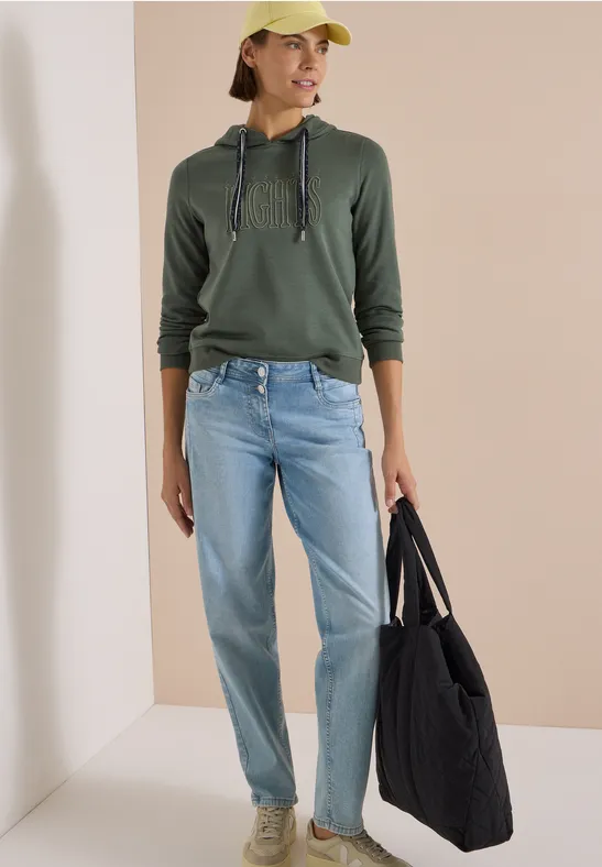 Eine Frau trägt grüne Kapuzenjacke, Jeans, beige Cap und hält eine große schwarze Tasche.
